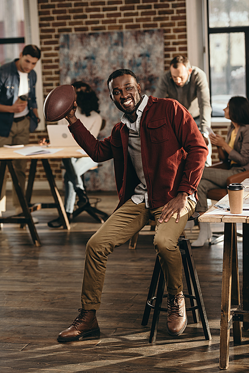 smiling african american casual businessman holding rugby ball with colleagues working behind in loft office
