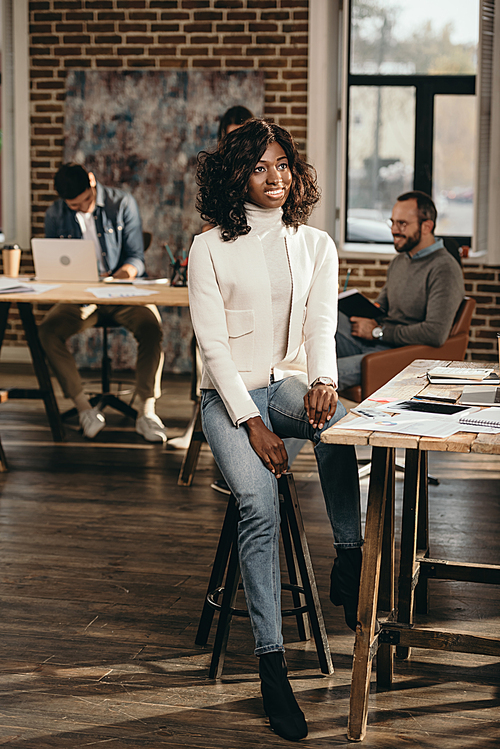 smiling african american casual businesswoman sitting in loft office with colleagues working behind