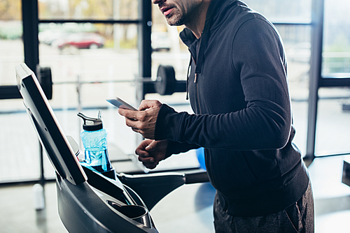 cropped image of sportsman in hoodie exercising on treadmill and using smartphone in gym