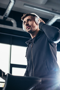 low angle view of handsome sportsman in hoodie exercising on treadmill and listening to music in gym