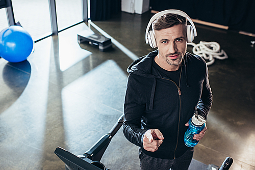 high angle view of handsome sportsman in hoodie standing on treadmill with sport bottle in gym