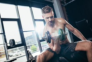 handsome muscular sportsman sitting on tire and exercising with in gym dumbbell