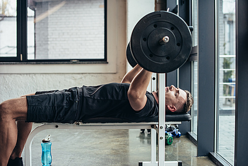 side view of handsome sportsman lifting barbell with weights while lying on bench in gym