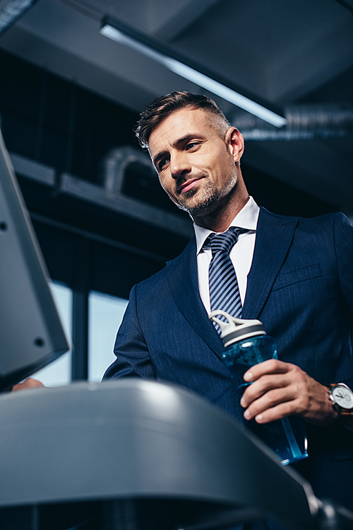 low angle view of handsome businessman in suit exercising on treadmill and holding sport bottle in gym