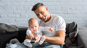 smiling father and baby daughter sitting on couch, holding remote controller and watching television at home