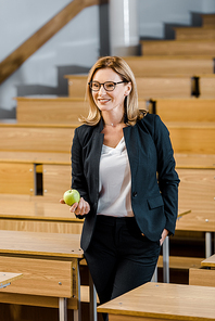 smiling female teacher in formal wear holding apple in classroom
