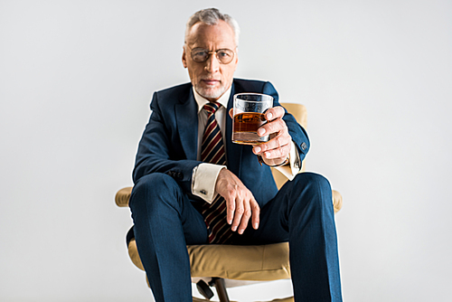 selective focus of mature businessman sitting in armchair and holding glass of whiskey isolated on grey