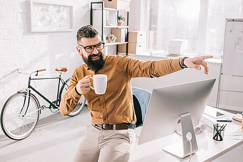 happy bearded adult businessman holding cup of coffee and pointing finger in exitement at workplace