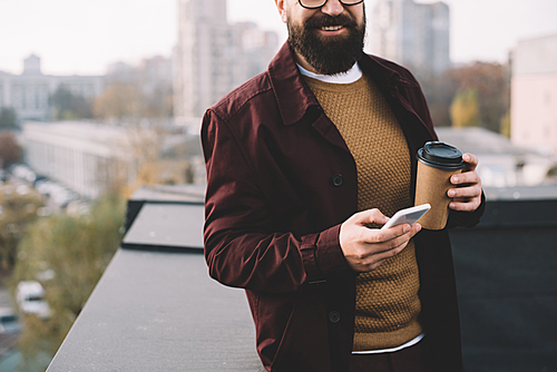 cropped view of stylish adult man in glasses holding coffee to go and using smartphone on rooftop