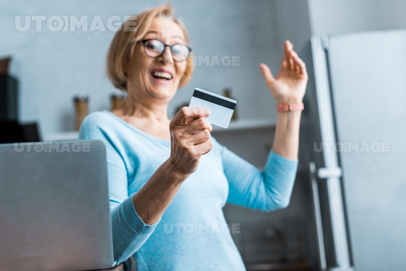 유토이미지 | excited senior woman in glasses gesturing with hand and holding credit card near laptop at home