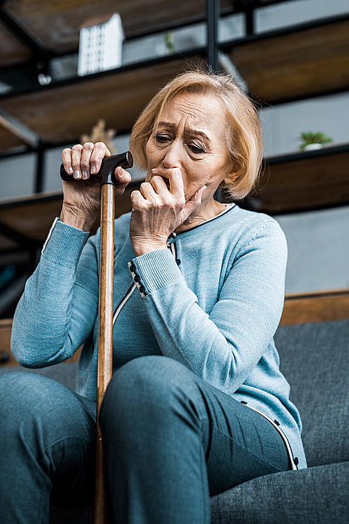depressed senior woman holding walking stick, covering mouth with hand and crying at home