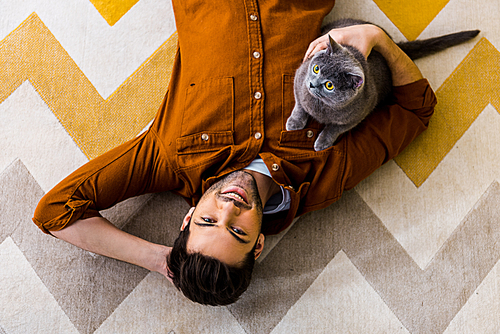 top view of smiling man lying on carpet with british shorthair cat