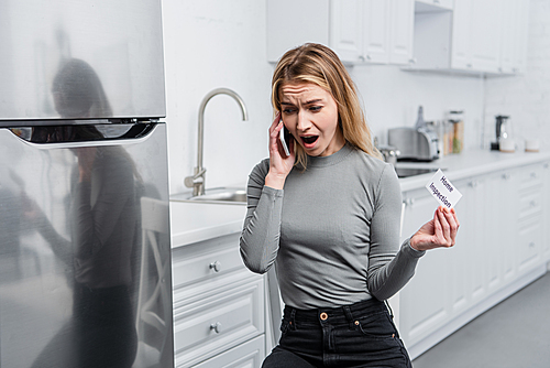 angry young woman holding card with lettering home inspection and yelling on smartphone in kitchen near broken refrigerator