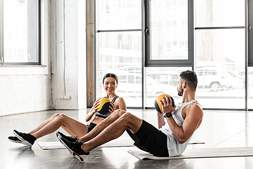 young couple holding medicine balls and smiling each other while training on yoga mats in gym