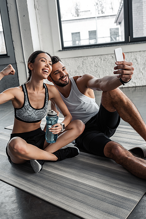 happy sporty young couple taking selfie with smartphone while resting on yoga mats after workout in gym