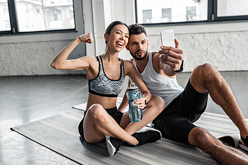 happy young couple in sportswear taking selfie with smartphone while resting on yoga mats after workout in gym