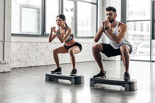 concentrated athletic young couple in sportswear squatting on step platforms in gym