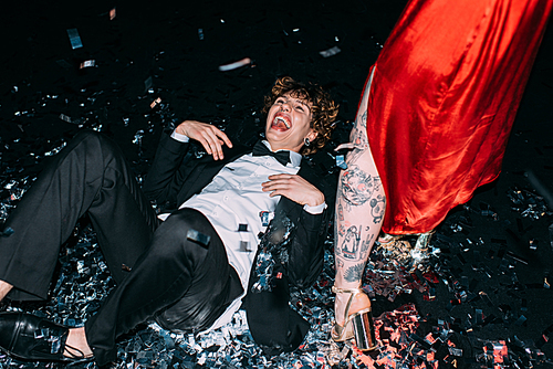 handsome man lying on floor with confetti and laughing near girlfriend isolated on black