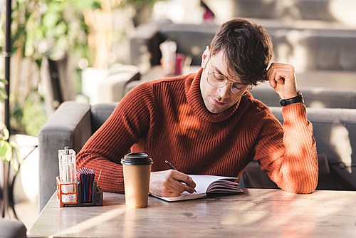 thoughtful man in glasses writing in notebook near disposable cup in cafe