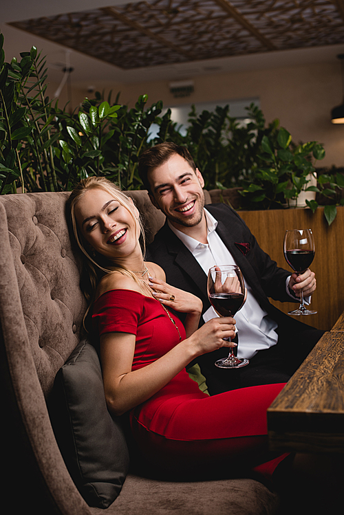 happy couple laughing while holding glasses with red wine in restaurant