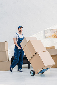 mover in uniform transporting cardboard boxes on hand truck in apartment with copy space