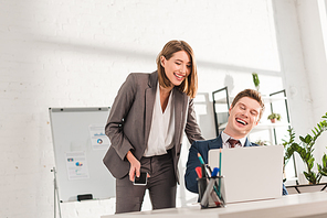 attractive businesswoman standing with smartphone near happy coworker and laughing while watching video on laptop