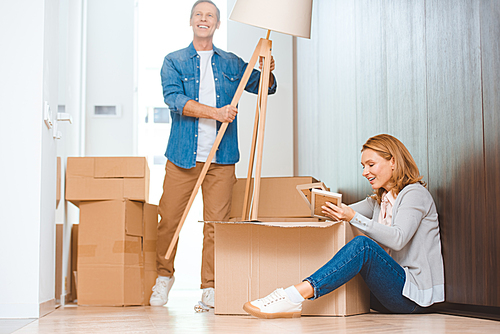 smiling man holding floor lamp while woman sitting in floor and unpacking carton box