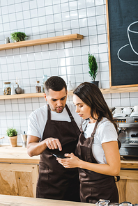 cashiers in brown aprons standing behind bar counter and looking to smartphone in coffee house