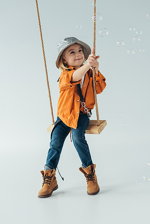 smiling kid in jeans and orange shirt sitting on swing and ponting with finger at soap bubbles