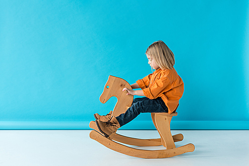 side view of blonde and cute kid riding on rocking horse on blue background