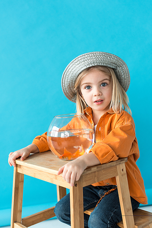 kid in silver hat and orange shirt sitting on stairs with fishbowl on blue background