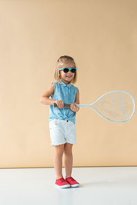 smiling and cute kid in sunglasses, shirt and shorts holding racket on beige background