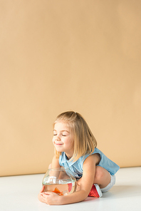 cute kid sitting with crossed legs and holding fishbowl on beige background