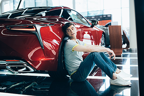 handsome man sitting near red car in car showroom