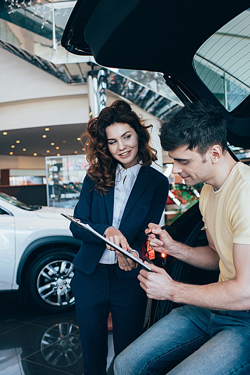 happy man signing paper near smiling car dealer in car showroom