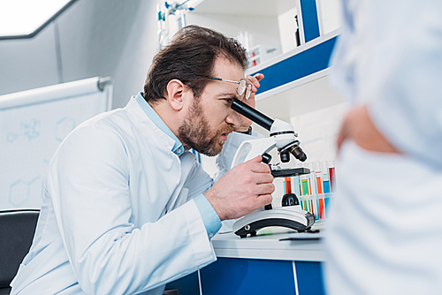 scientist in lab coat looking through microscope on reagent with colleague near by in laboratory