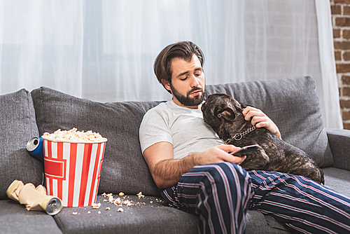 handsome loner palming bulldog and holding remote control on sofa in living room