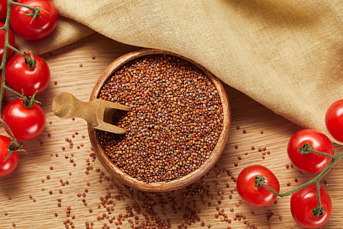 top view of red quinoa in wooden bowl with spatula near beige napkin and tomatoes