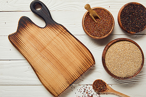 top view of white, black and red quinoa in wooden bowls near chopping board on white table