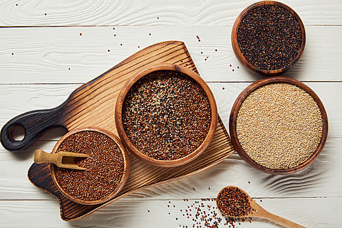 top view of white, black and red quinoa in wooden bowls on white table with chopping board