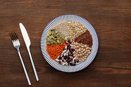 top view of fork and knife near striped plate with raw lentil, chickpea, quinoa, oatmeal, beans and pumpkin seeds on wooden surface
