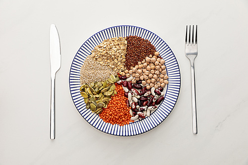 top view of striped plate with raw lentil, chickpea, quinoa, oatmeal, beans and pumpkin seeds near cutlery on marble surface