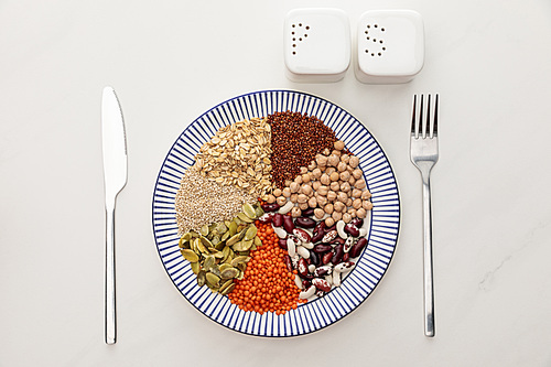 top view of plate with raw cereal, diverse beans and pumpkin seeds near cutlery and salt and pepper pots on marble surface