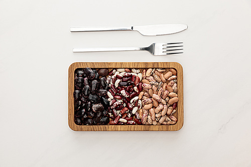 top view of rectangular wooden dish with raw beans near fork and knife on white marble surface