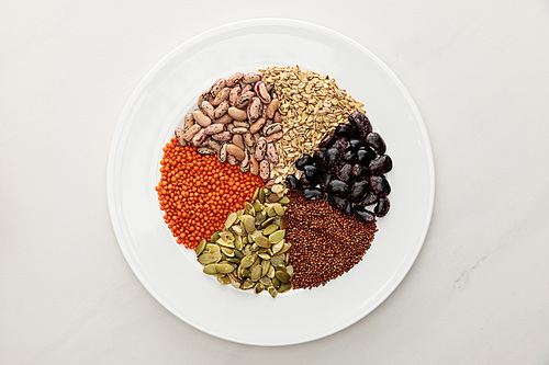 top view of white plate with raw lentil, quinoa, oatmeal, beans and pumpkin seeds on marble surface