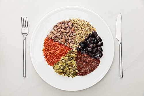 top view of white plate with raw lentil, quinoa, oatmeal, beans and pumpkin seeds near fork and knife on marble surface