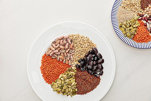 top view of white and striped plates with raw lentil, quinoa, oatmeal, beans and pumpkin seeds on marble surface
