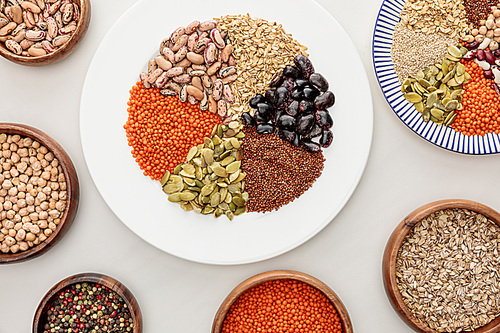 top view of white and striped plates and wooden bowls with raw lentil, quinoa, oatmeal, beans, peppercorns and pumpkin seeds on marble surface