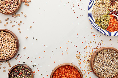 top view of plate and bowls with raw lentil, quinoa, oatmeal, beans, peppercorns and pumpkin seeds on marble surface with scattered grains and copy space