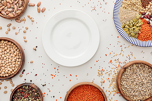 top view of empty plate and bowls with raw lentil, quinoa, oatmeal, beans, peppercorns and pumpkin seeds on marble surface with scattered grains